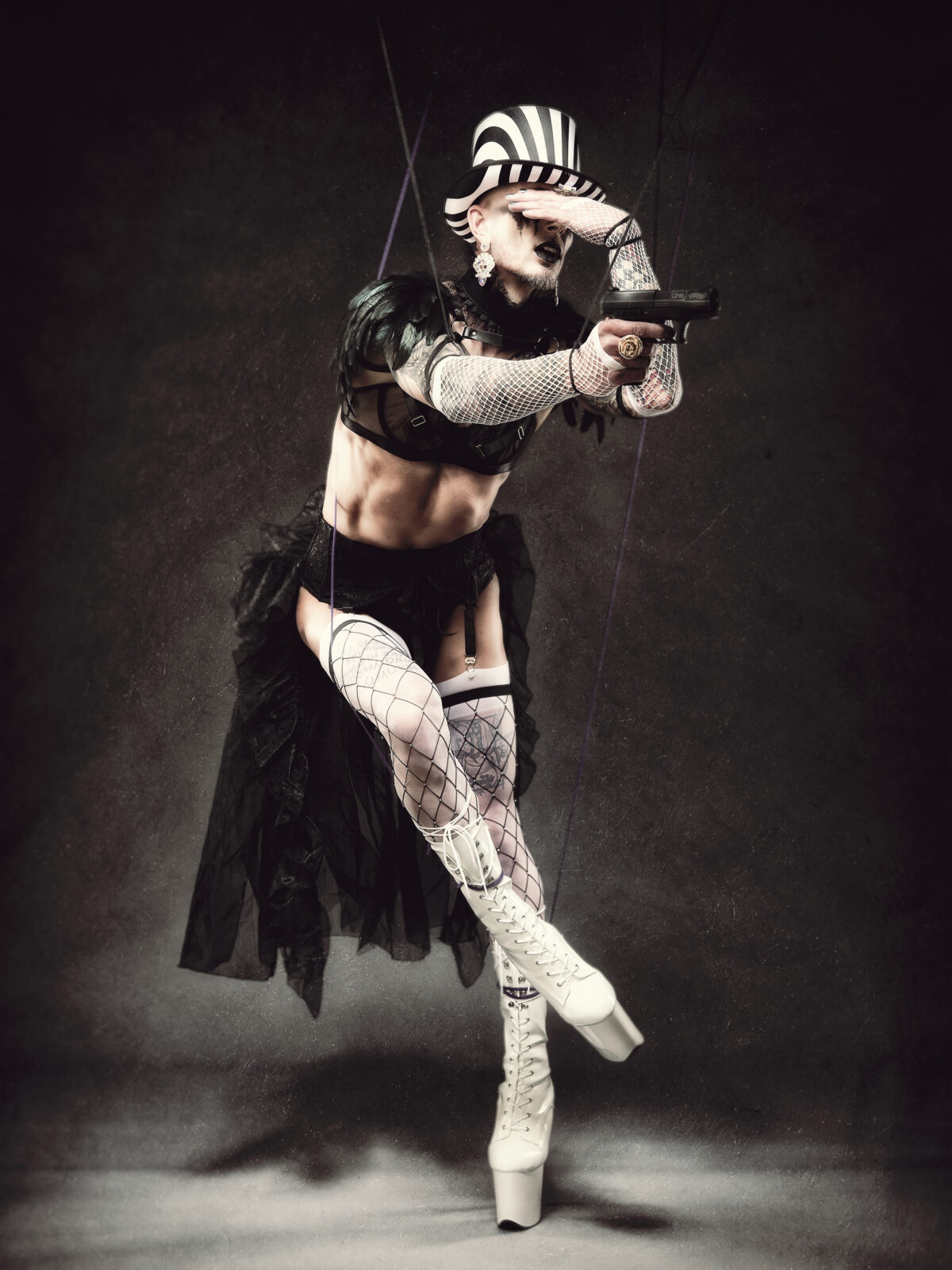 Queeres Studiofoto einer Marionettenfigur mit Netzstrümpfen, Lackmode und Pistole – Fetischfotograf Hell On Heels Photography in Düsseldorf und Köln